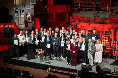 Many people stand on the stage in front of the blast furnace in the DASA steel hall.