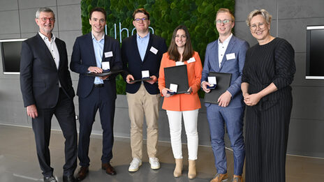 Six people stand in a row next to each other, the middle four holding a folder and a box with a medal in front of them.