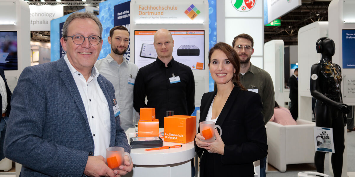 Mehrere Personen stehen an einem Tisch, im Hintergrund ist ein Messestand mit dem Wappen von Nordrhein-Westfalen zu sehen.