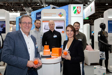 Several people are standing at a table, in the background you can see an exhibition stand with the coat of arms of North Rhine-Westphalia.