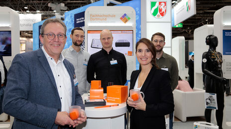 Several people are standing at a table, in the background you can see an exhibition stand with the coat of arms of North Rhine-Westphalia.