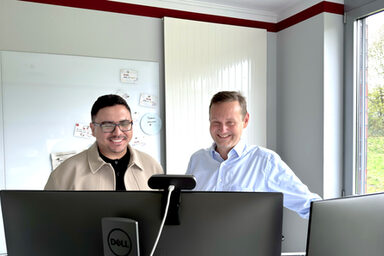 Two people stand behind two monitors and look at them.