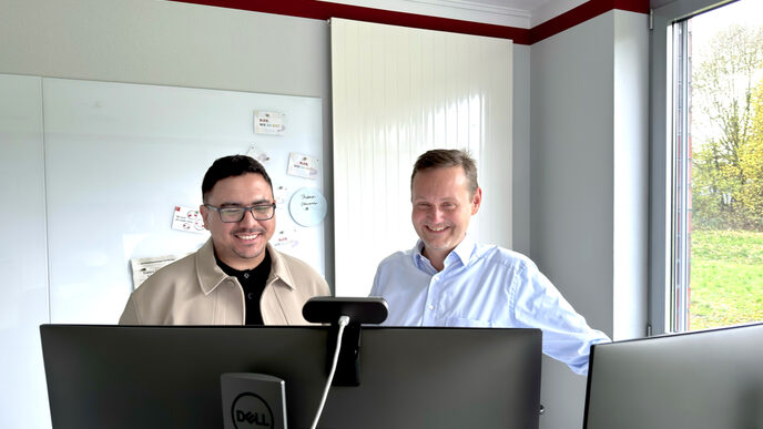 Zwei Personen stehen hinter zwei Monitoren und blicken darauf.