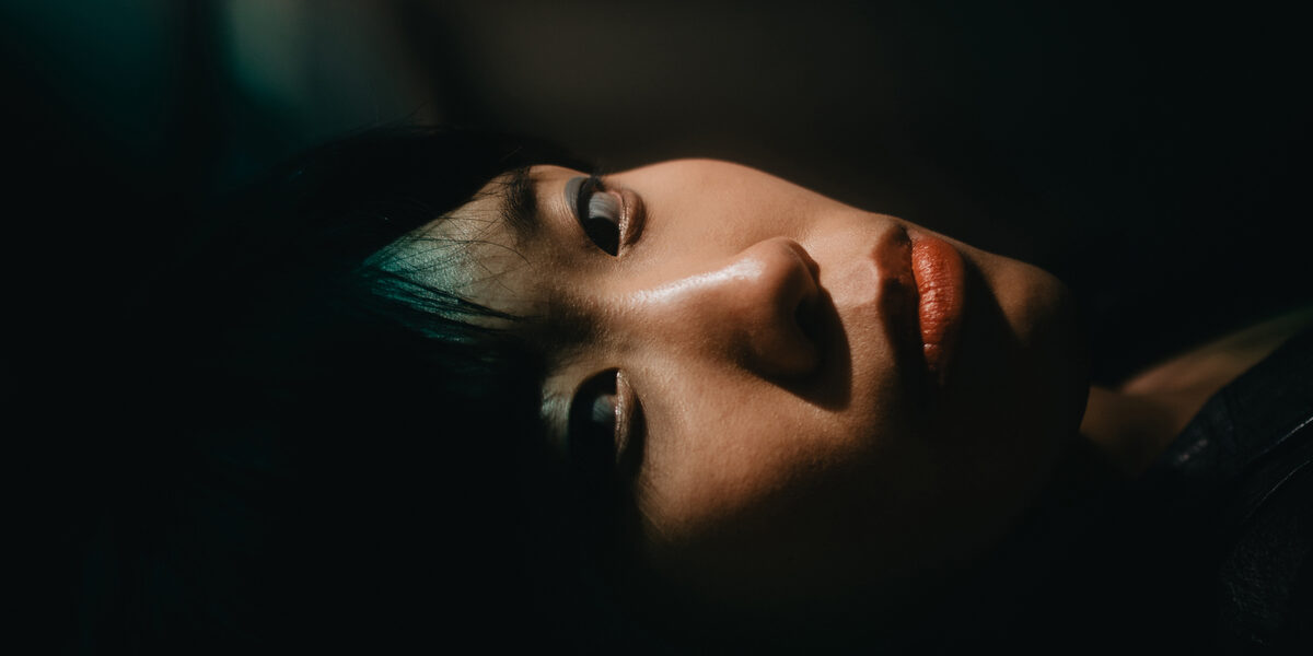 The head of a person lying on his side, his face and gaze directed towards the camera, against a dark background and a dark green background.