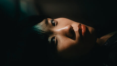 The head of a person lying on his side, his face and gaze directed towards the camera, against a dark background and a dark green background.