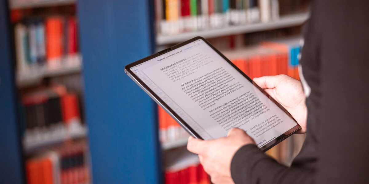 Foto von einem Tablet in der Hand von jemanden, der in einer Bibliothek steht.