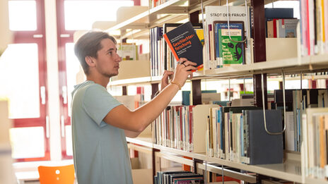 Foto von einem Studierenden, der ein Buch aus einem Bücherregal der Bibliothek holt.