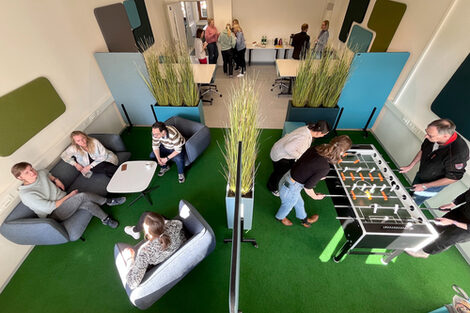 Foto von mehreren Personen, die sich in der BibLounge am Standort Sonnenstraße aufhalten. Eine Gruppe unterhält sich am Eingang, eine weitere sitzt auf Sesseln und einem Sofa und eine dritte Gruppe spielt Tischfußball__Photo of three groups of people in the BibLounge at campus Sonnenstraße. One group is standing by the entrance, another is sitting in armchairs and a sofa, and the third one is playing table football.