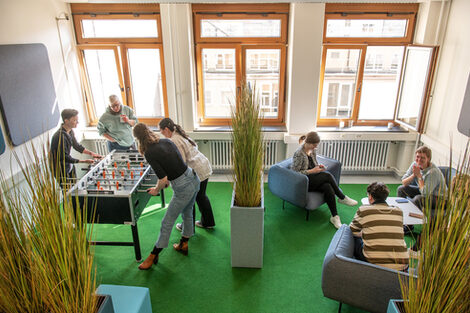 Foto von mehreren Personen, die sich in der BibLounge am Standort Sonnenstraße aufhalten. Eine Gruppe sitzt auf Sesseln und einem Sofa, eine weitere Gruppe spielt Tischfußball__Photo of two groups of people in the BibLounge at campus Sonnenstraße. One group is sitting in armchairs and a sofa, the other one is playing table football.