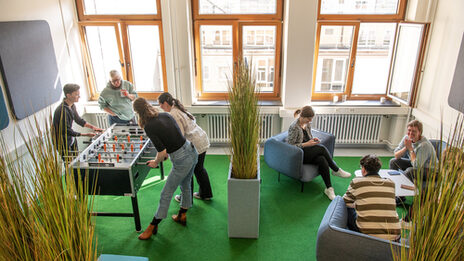 Foto von mehreren Personen, die sich in der BibLounge am Standort Sonnenstraße aufhalten. Eine Gruppe sitzt auf Sesseln und einem Sofa, eine weitere Gruppe spielt Tischfußball__Photo of two groups of people in the BibLounge at campus Sonnenstraße. One group is sitting in armchairs and a sofa, the other one is playing table football.