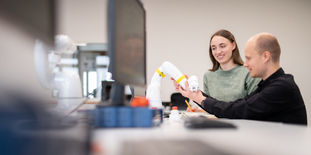 Foto von zwei Personen, die sich mit einem Schwenkarmroboter beschäftigen. Eine Person schaut sich den Schrenkarmroboter an, während die andere Person auf dem Bild den Arm bedient.