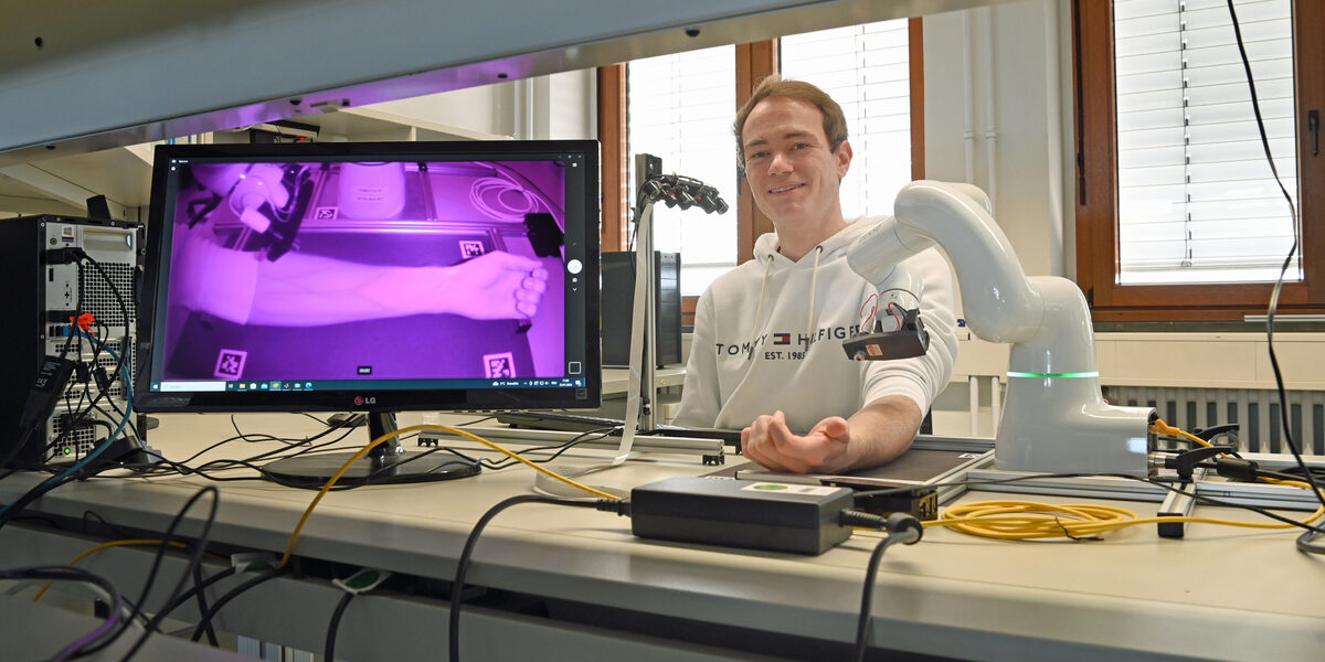 Eine Person sitzt an einem Labor-Tisch und hat den Unterarm unter einen Roboter gelegt. Auf einem Bildschirm daneben werden die Venen des Unterarms im Infrarotlicht deutlich sichtbar.