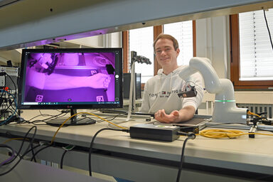 A person is sitting at a laboratory table and has placed their forearm under a robot. The veins of the forearm are clearly visible in infrared light on a screen next to it.