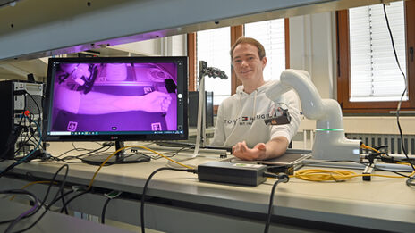 A person is sitting at a laboratory table and has placed their forearm under a robot. The veins of the forearm are clearly visible in infrared light on a screen next to it.