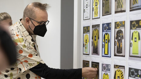 Ein Mann mit Maske steht vor einer Wand mit mehren Bildern von Trikots und markiert eines dieser Bilder mit einem Klebepunkt.