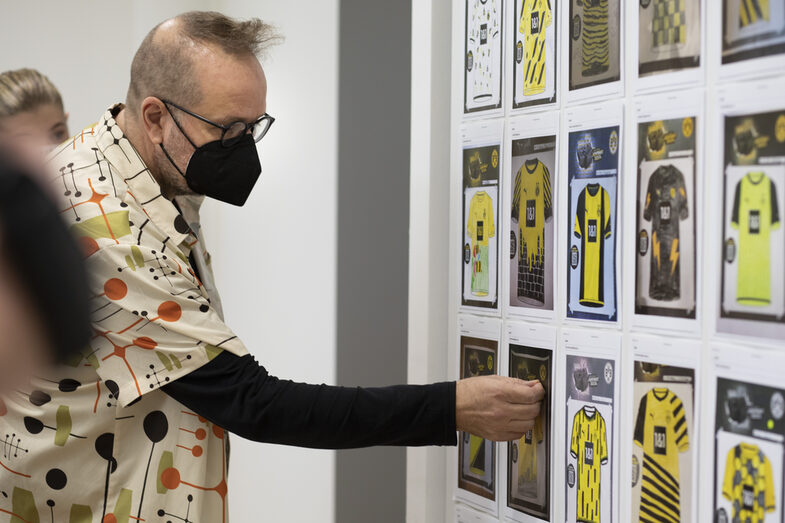 Ein Mann mit Maske steht vor einer Wand mit mehren Bildern von Trikots und markiert eines dieser Bilder mit einem Klebepunkt.