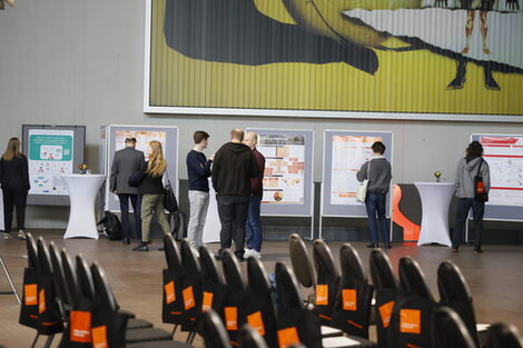 Im Vordergrund sind Stühle zu sehen. Im Hintergrund schauen sich die mehrere Teilnehmenden interessiert die auf großen Pinnwänden ausgestellten Poster der Promovierenden an.