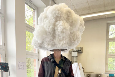 Eine Person steht unter einem wolkenartigen Gebilde, das unten eine Öffnung aufweist, die den Kopf der Person verdeckt.