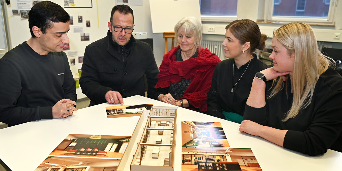 A group of people sits around a model and illustrations and discusses the designs.