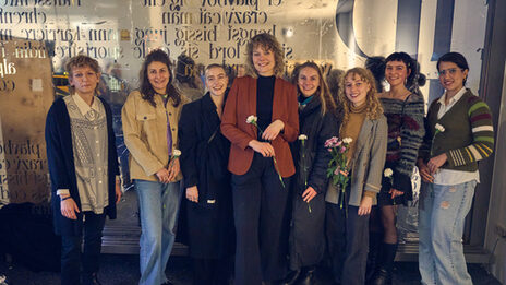 Eight people stand next to each other in front of a shop window covered with lettering and look friendly into the camera.