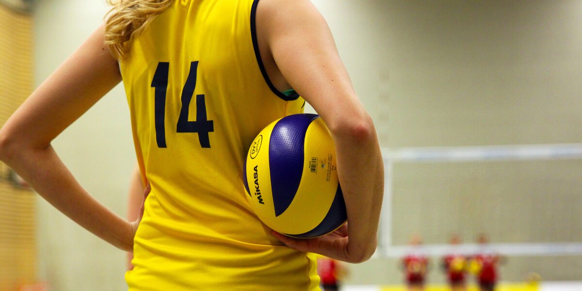 A person read as a female is standing on a volleyball court in a sports hall, holding a ball under her arm.