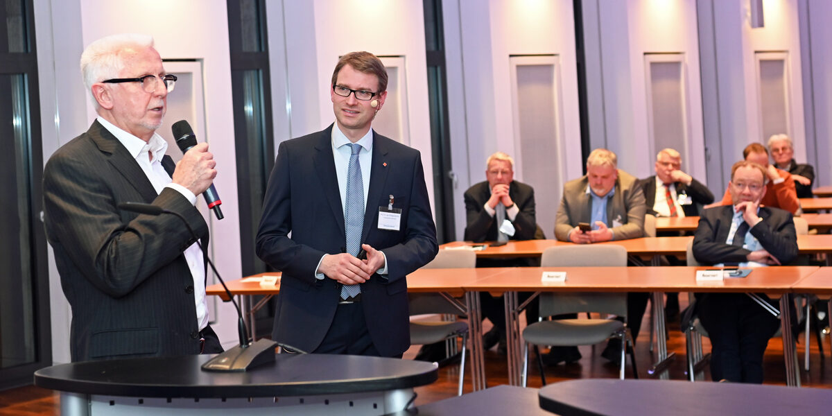 Two people are standing at a lectern and talking. The audience can be seen in the background.