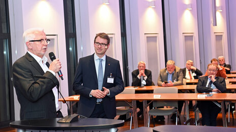 Two people are standing at a lectern and talking. The audience can be seen in the background.