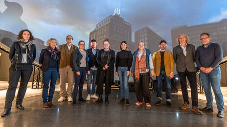 Gruppenbild mit 11 Personen, die nebeneinander vor einer Projektion stehen, die das Dortmunder U von außen zeigt.
