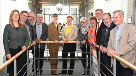 Eleven people stand around the banister of a staircase and look into the camera.