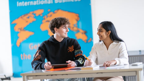 Foto von zwei Studierenden, die an einem Tisch sitzen und sich unterhalten. Vor ihnen auf dem Tisch liegen Arbeitsmaterialien. An der Wand hinter den Beiden hängt eine orangene Weltkarte auf blauem Grund mit der Überschrift „International Business“.