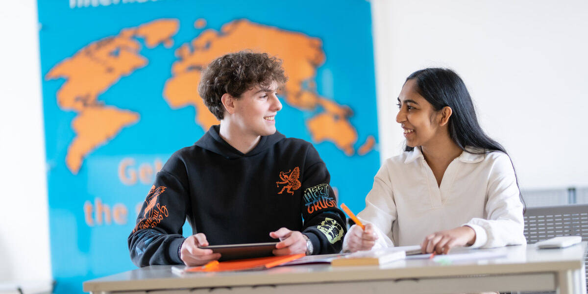 Photo of two students sitting at a table and talking. In front of them on the table are work materials. On the wall behind them hangs an orange world map on a blue background with the heading "International Business".