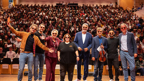 Mehre Personen mit Sonnenbrille stehen auf einer Bühne. Im Hintergrund ist ein voller Saal zu sehen.