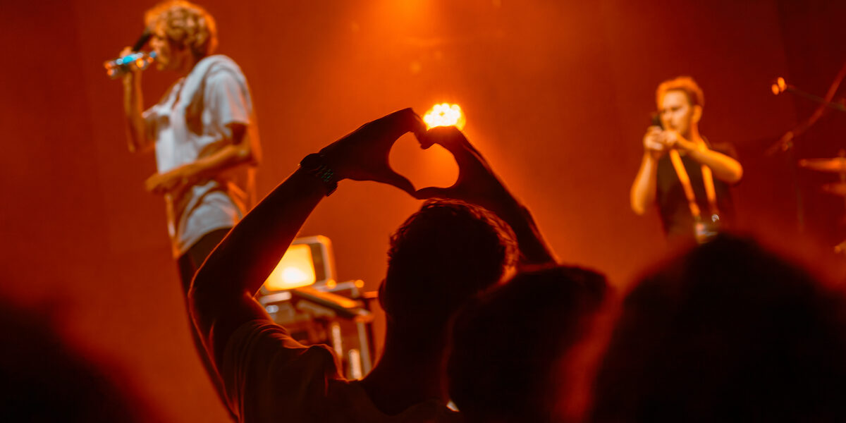 In the foreground, a person forms a heart with his hands; in the background, musicians can be seen on a stage.