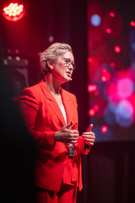 A person in a red suit stands on a colorfully lit stage and speaks to an audience.