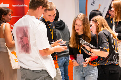 Several young people are looking together at a cell phone that one of them is holding out to the others.