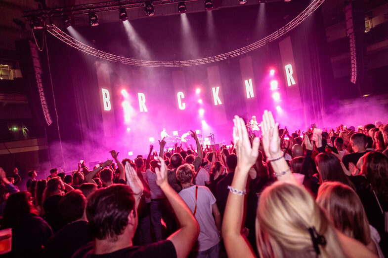 Blick aus dem Publikum auf die Bühne: Erhobene, klatschende Hände im Vordergrund, die violett beleuchtete Bühne mit dem Schriftzug "BRCKNR" im Hintergrund.