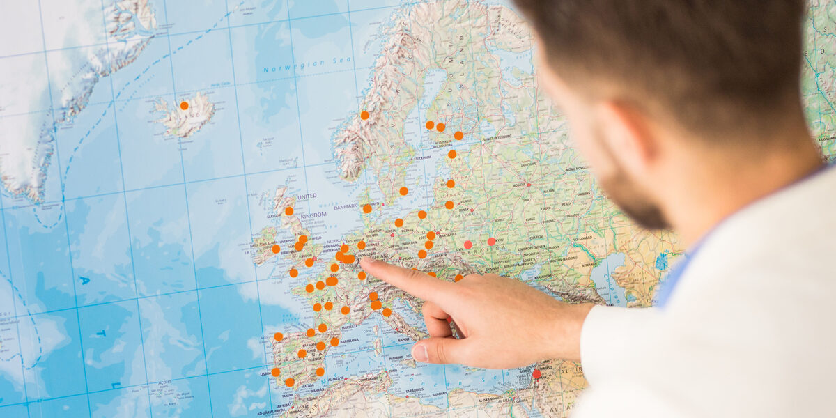 Foto einer Person, die auf eine große Weltkarte zeigt, auf der mit orangefarbenen Punkten Orte markiert sind