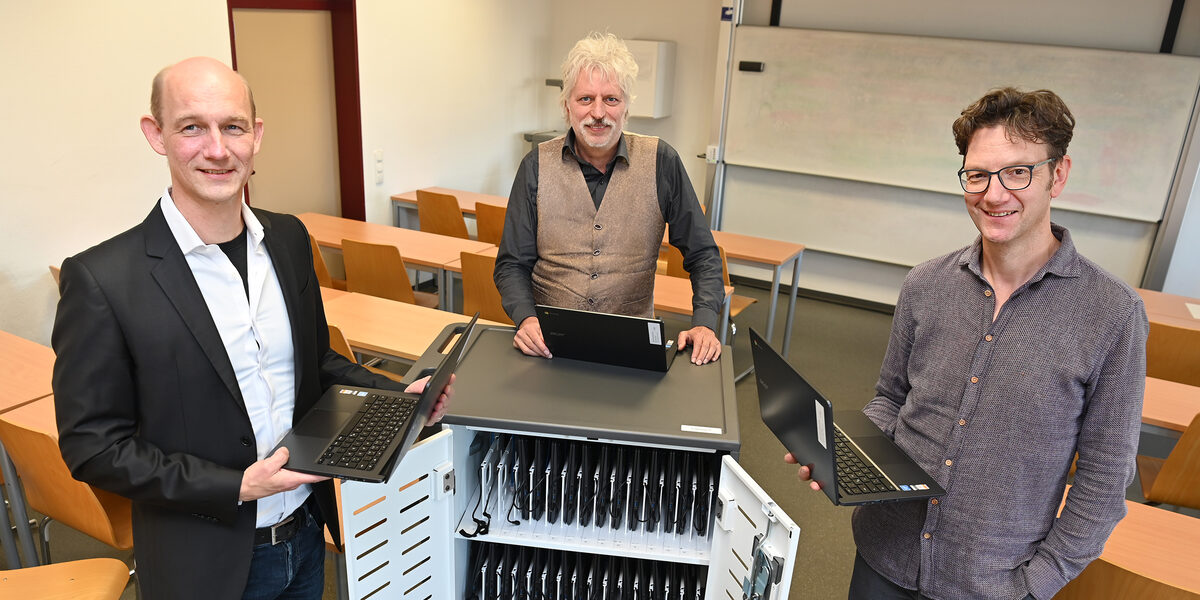 Three people are standing around a roll container filled with notebooks. Two of them are holding a laptop.