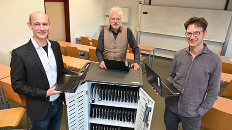 Drei Personen stehen um einen Rollcontainer der mit Notebooks bestückt ist. Zwei von ihnen halten einen Laptop in der Hand.