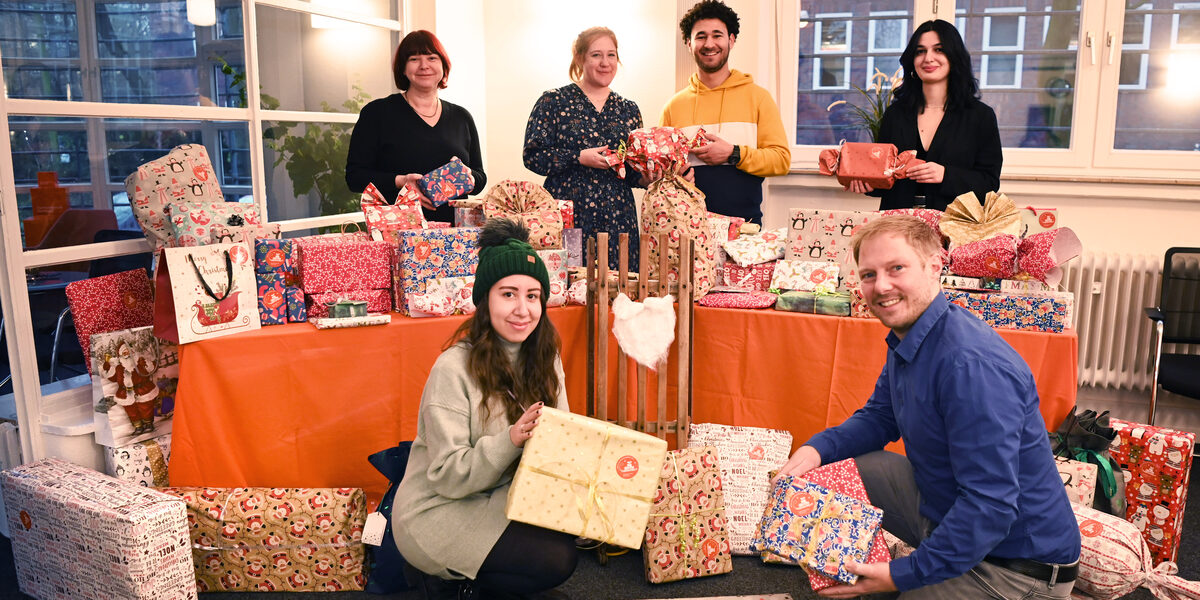 Four people are standing behind a table covered in orange with lots of wrapped presents, each holding presents in their hands. Two more people are sitting in front of the table. They are all smiling and looking at the camera.