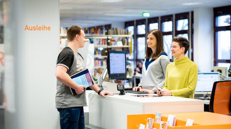 Foto von zwei Mitarbeiterinnen, die hinter der Theke der Bibliothek stehen und mit dem ihnen gegenüber stehenden Studierenden, der Bücher in der Hand hat, im Gespräch sind.