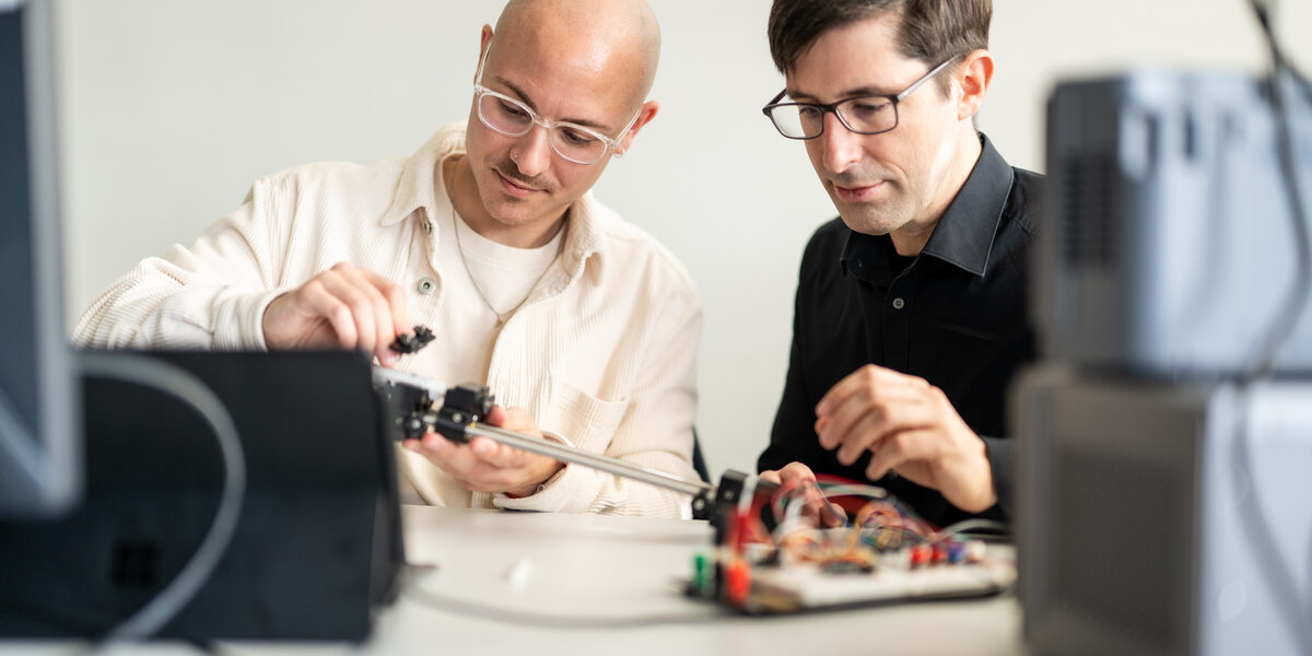 Bild von einem Studierenden neben seinem Professor. Beide sitzen an einem Versuchsaufbau.