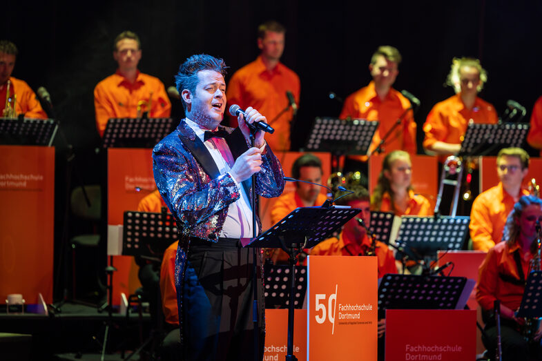 Ein Sänger mit glitzerndem Jacket singt. Im Hintergrund ist die Band in orangefarbenen Hemden zu sehen.