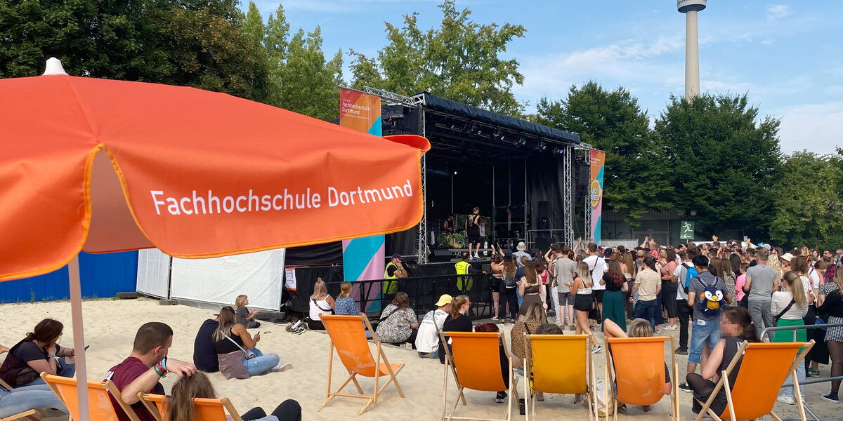 Menschen sitzen in den FH-Dortmund-Liegen unter den Sonnenschirmen vor einer Bühne.