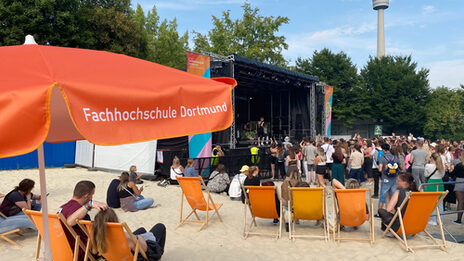 Menschen sitzen in den FH-Dortmund-Liegen unter den Sonnenschirmen vor einer Bühne.