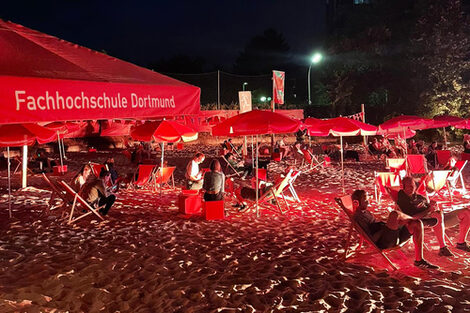 Unter den Sonnenschirmen der FH sitzen Menschen nachts auf den Liegen am Strand beim Juicy-Beats-Festival.