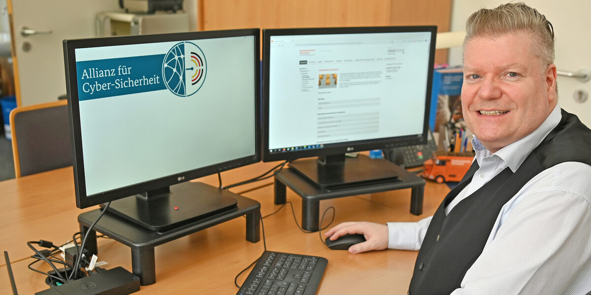 Eine männlich gelesene Person sitzt vor zwei Monitoren am Schreibtisch. Auf einem Monitor ist das Logo der Allianz für Cyber-Sicherheit zu sehen.