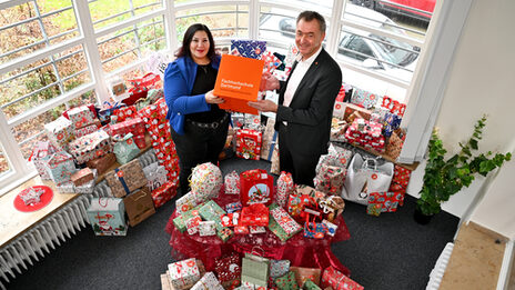 Zwei Personen stehen inmitten von weihnachtlich verpackten Geschenken und halten gemeinsam einen orangefarbenen FH-Dortmund-Würfel in den Händen.