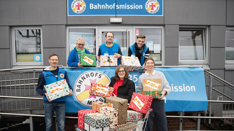 Six people stand at the entrance to the Dortmund station mission. Each of them is holding a festively wrapped parcel in their hands. In the middle in front is a shopping cart full of more gift packages.