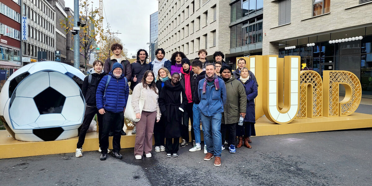 18 people stand on a square between multi-storey buildings in front of a three-dimensional "Dortmund" lettering with an armchair in the shape of an oversized soccer.